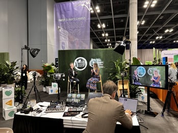 A media studio setup at the NEST Climate Campus, showing a man and a woman having a conversation on stage in front of a green backdrop with plants and event signage. In the foreground, a producer operates audio and video equipment, while a large monitor displays a live feed of the speakers. The scene is lively and professional, reflecting climate-focused discussions at a conference.