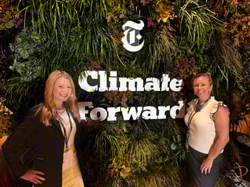 Two women (Kim Lundgren on the right) smiling and standing in front of a lush green wall decorated with foliage and the New York Times “Climate Forward” event sign. Both are wearing event badges, posing in a well-lit, professional event setting.