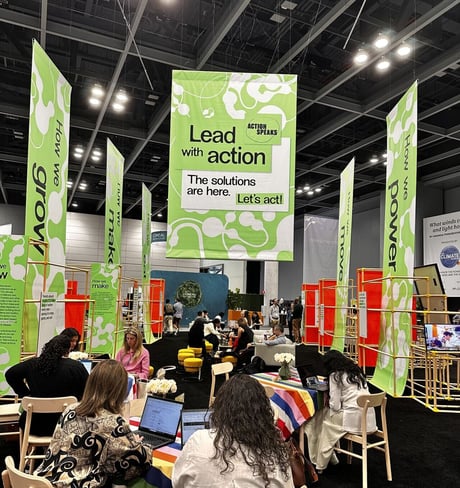 People work and converse at tables covered with colorful striped tablecloths in a large convention hall. Tall green banners hang from the ceiling and display climate action messages such as ‘Lead with action. The solutions are here. Let’s act!’ The space is lively with groups talking, laptops open, and event signage focused on growth, power, and making change.