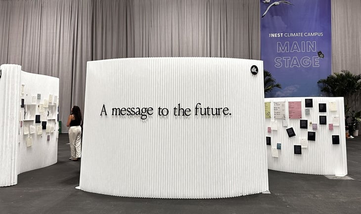 Exhibit display at the NEST Climate Campus featuring a large white wall with black text that reads “A message to the future.” In the background, additional curved white walls are covered with colorful handwritten notes, and a blue banner for the Main Stage hangs overhead. A person stands reading the notes, emphasizing community engagement and climate-focused reflection.